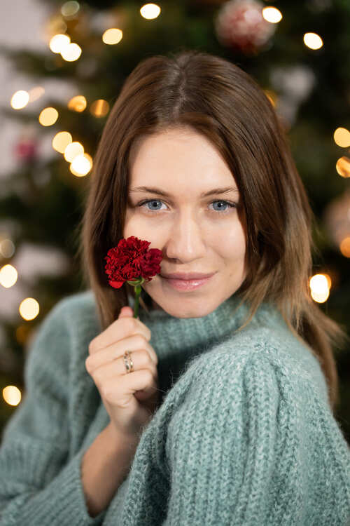 Ritratto di donna che tiene un fiore rosso davanti al viso con albero di Natale illuminato sullo sfondo