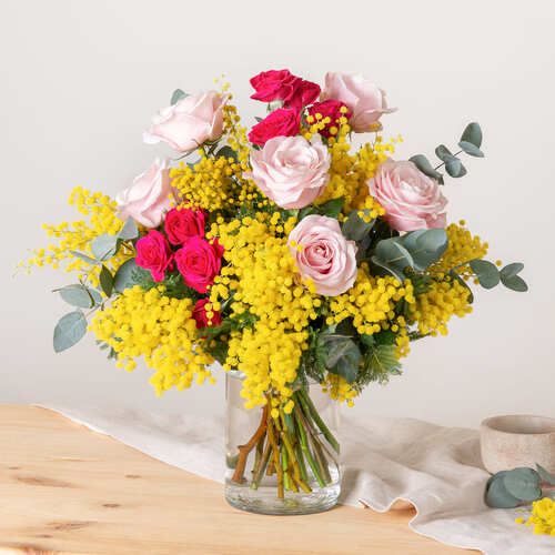 Bouquet misto con mimosa gialla, rose rosa e fucsia ed eucalipto, sistemato in vaso di vetro su tavolo in legno