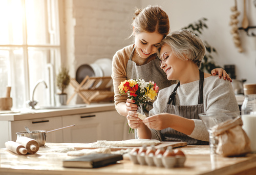 Figlia adulta regala un piccolo bouquet alla madre mentre trascorrono un momento insieme in cucina preparando dolci.